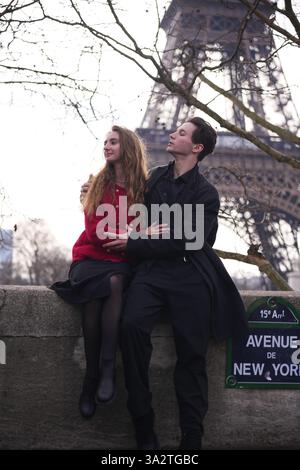 Un jeune couple partage un moment romantique sur l'avenue de New York à Paris, assis sur une corniche de pierre avec la Tour Eiffel derrière eux. Banque D'Images