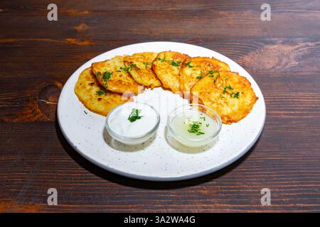 Des crêpes de pommes de terre fraîchement préparées reposent sur une assiette blanche, accompagnées de deux petits bols de trempettes. Le cadre dispose d'une table en bois rustique, mettant en valeur le A. Banque D'Images