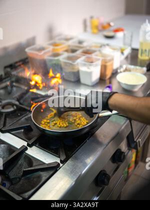 Cuisinier professionnel portant des gants noirs, ajoutant des pois chiches dans une poêle remplie d'épices aromatiques, préparant habilement la cuisine indienne traditionnelle dans un Banque D'Images