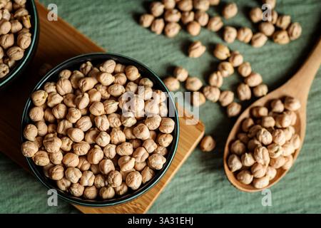 Gros plan d'un bol en céramique rempli de pois chiches séchés sur un plateau en bois avec des légumineuses éparpillées sur un fond vert texturé, mettant en évidence fo naturel Banque D'Images
