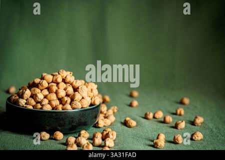 Gros plan d'un bol en céramique noire rempli de pois chiches crus, placé sur un tissu vert avec des légumineuses éparpillées autour. La lumière naturelle met en valeur leur Banque D'Images