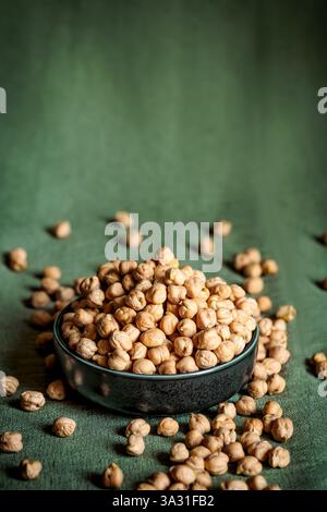 Bol en céramique noire rempli de pois chiches crus, entouré de légumineuses éparpillées sur un tissu vert. Un éclairage chaud et naturel rehausse le rustique et l'organi Banque D'Images