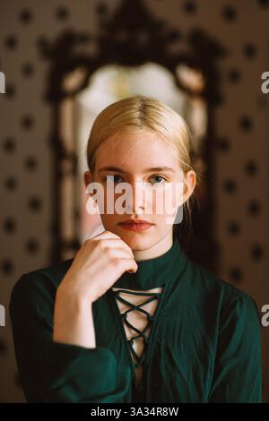 Une jeune femme tranquille aux cheveux blonds et aux yeux bleus porte une robe à lacets vert foncé, regardant avec confiance la caméra. Un miroir vintage et une douceur de vivre Banque D'Images