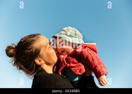 Une mère portant un pull foncé embrasse son bébé, vêtu d'un pull rouge et d'un chapeau vert, contre un ciel bleu vif. Le moment tendre capte la chaleur Banque D'Images