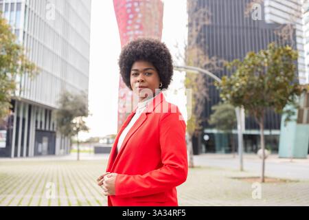 Une femme confiante dans une veste rouge pose dans un cadre urbain moderne. Sa coiffure afro contraste magnifiquement avec l'arrière-plan architectural élégant. Banque D'Images