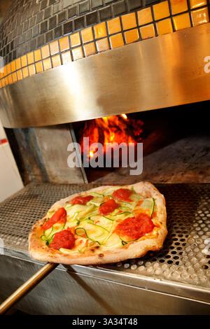 Une pizza fraîchement préparée avec des garnitures de tomate, pepperoni et tranches de courgettes est cuite dans un four en briques au feu de bois incandescent, mettant en valeur l'art. Banque D'Images