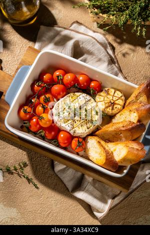 Dégustez un élégant plat de camembert cuit au four accompagné de tomates cerises, d'ail rôti et de tranches de baguette grillées, garni d'herbes fraîches pour Banque D'Images