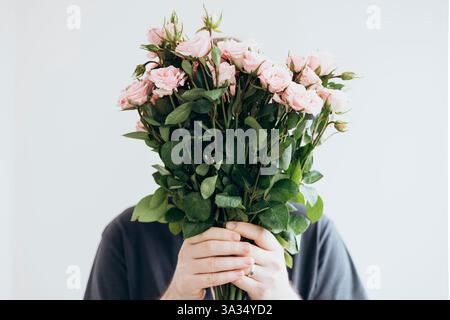 Un jeune homme tient un bouquet de roses roses roses douces, partiellement cachées derrière les fleurs. Le fond neutre rehausse les pétales délicats et le vert luxuriant Banque D'Images