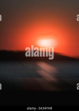 Une image abstraite et floue capturant un coucher de soleil rouge éclatant sur un océan calme. La distorsion crée un sentiment de mouvement et une atmosphère onirique, parfait Banque D'Images