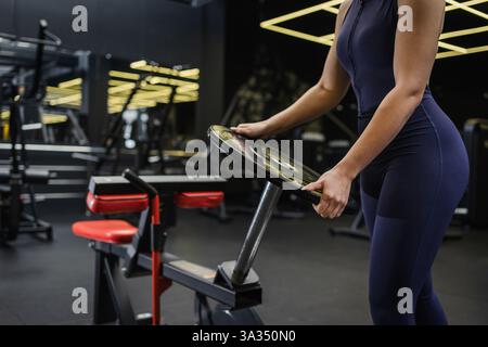 Une femme en sportswear sombre tient une plaque de poids dans un cadre de gymnastique moderne. Le centre de fitness est équipé d'appareils d'entraînement de pointe et d'un intérieur élégant Banque D'Images