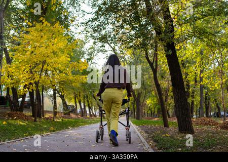 Une femme atteinte de paralysie cérébrale se promène avec un marcheur sur un sentier pittoresque entouré d'arbres d'automne vibrants. Le feuillage coloré crée un serein et pe Banque D'Images
