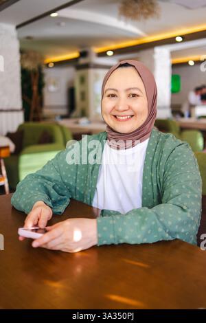 Une femme musulmane souriante dans un hijab s'assoit confortablement à une table à l'intérieur, tenant un smartphone. Elle respire la chaleur et la positivité, soulignant d culturel Banque D'Images