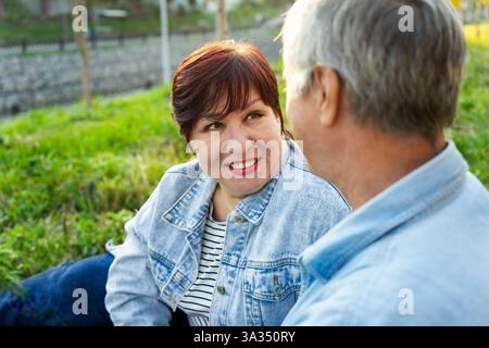 Un couple aîné assis près les uns des autres sur un champ herbeux, partageant une conversation joyeuse. La femme sourit chaleureusement, portant une veste en denim et une bande Banque D'Images