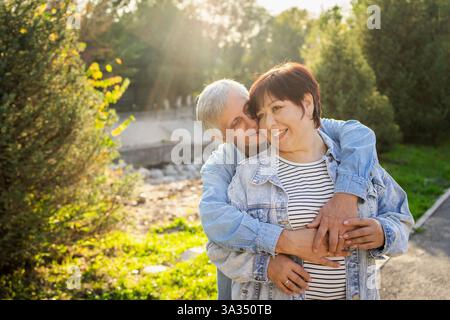 Un couple âgé partage une étreinte aimante, vêtu de façon décontractée, avec des sourires sur leurs visages. Ils profitent d'une journée ensoleillée dans un parc verdoyant luxuriant, se prélasser dans la chaleur Banque D'Images