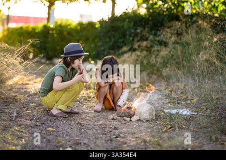 Deux enfants, dont un en chapeau, s’assoient près d’un petit feu de camp dans un cadre naturel. Entourés d'arbres et d'herbe, ils observent attentivement les flammes, expérienci Banque D'Images