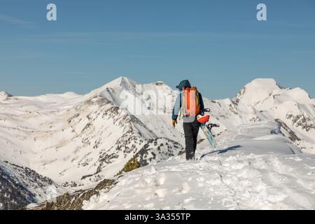 Vue arrière d'une personne méconnaissable qui grimpe une montagne enneigée avec un snowboard. Le snowboarder, vêtu de vêtements d'hiver, est entouré d'un Banque D'Images