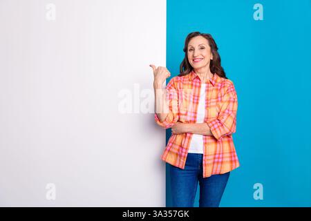 Une femme âgée heureuse portant une chemise à carreaux sourit et pointe avec confiance sur un fond élégant à deux tons, exsudant charme et positivité. Banque D'Images