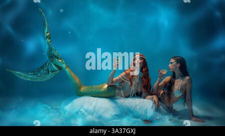 Moment serein en haute mer entre deux gracieuses sirènes dégustant du champagne contre un océan bleu profond mystique. Banque D'Images
