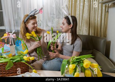 Une mère et sa fille portant des oreilles de lapin partagent un moment joyeux tout en tenant des tulipes jaunes dans un cadre confortable de Pâques. Banque D'Images