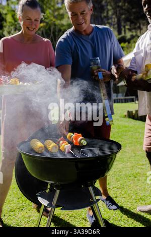 Divers amis seniors appréciant barbecue en plein air, griller des légumes et socialiser dans la cour arrière Banque D'Images