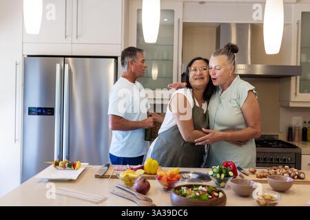Dans la cuisine, les femmes âgées embrassant tandis que l'homme prépare le repas avec des légumes frais Banque D'Images