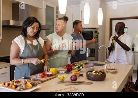 Dans la cuisine moderne, des amis seniors préparant des brochettes de légumes et buvant ensemble Banque D'Images