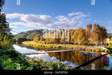 Une rivière calme coule sous un pont suspendu entouré d'arbres d'automne vibrants dans le centre de Lyngdal dans le sud de la Norvège, reflet du ciel bleu Banque D'Images