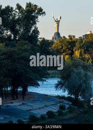Au milieu d'une végétation luxuriante, l'imposant Monument de mère Ukraine se dresse sentinelle au bord du fleuve Dnipro alors que le crépuscule jette une lueur dorée sur Kiev. Banque D'Images