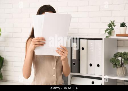 Jeune femme cachant son visage derrière des documents au bureau. Gros plan Banque D'Images
