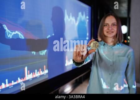Gros plan de pièce de monnaie numérique en main. Femme d'affaires est contre projecteur avec des graphiques, à l'intérieur. Banque D'Images