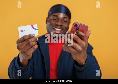 Carte de crédit et smartphone dans les mains, effectuer le paiement. Bel homme noir est dans le studio sur fond jaune. Banque D'Images
