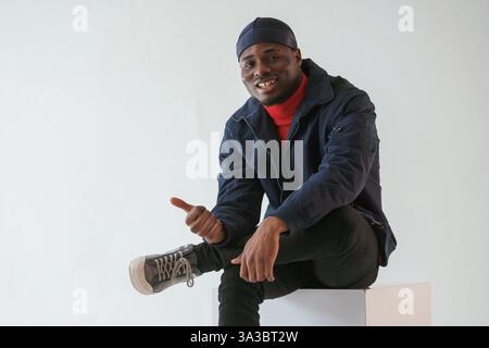 Calme, assis, souriant et montrant le pouce vers le haut. L'homme noir en casquette est dans le studio sur fond blanc. Banque D'Images