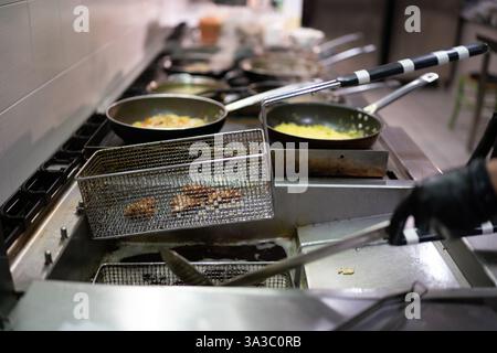Chef professionnel faisant frire pakora dans un panier immergé dans l'huile bouillante dans une cuisine animée, en utilisant un wok et une poêle à frire pour préparer authentique Banque D'Images