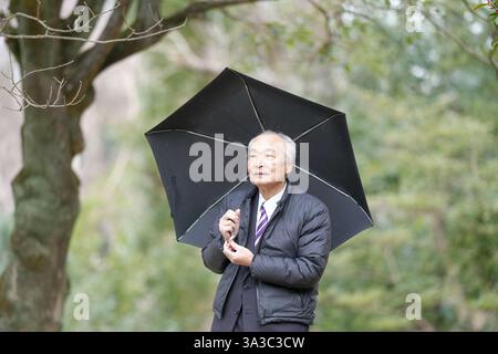 Un parc un jour de pluie au Japon avec beaucoup de verdure au printemps. Un Japonais à la fin des années 70 est debout dans un costume et une veste en duvet. Il pointe vers t Banque D'Images