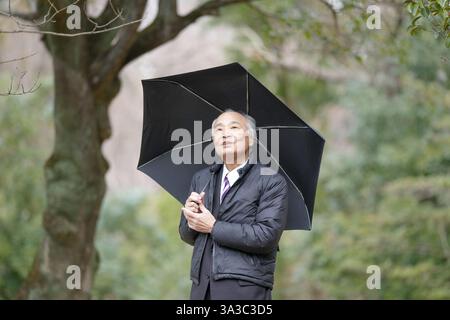 Un parc un jour de pluie au Japon avec beaucoup de verdure au printemps. Un Japonais à la fin des années 70 est debout dans un costume et une veste en duvet. Il pointe vers t Banque D'Images