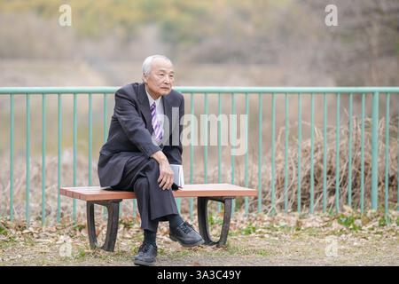 Un parc de jour de printemps confortable avec beaucoup de verdure au Japon. Un Japonais à la fin des années 70 est assis sur un banc portant un costume. Il tient un bo Banque D'Images