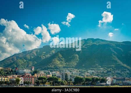 Colorful houses nestled at the foothills of lush green mountains on the Amalfi Coast in Italy, under a vibrant blue sky dotted with white clouds, show Banque D'Images