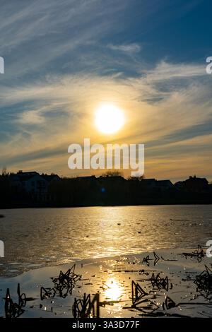Un coucher de soleil serein jette une lueur chaude sur un lac calme, illuminant la surface de l'eau. Des silhouettes de maisons bordent l'horizon contre un beau ciel. Banque D'Images