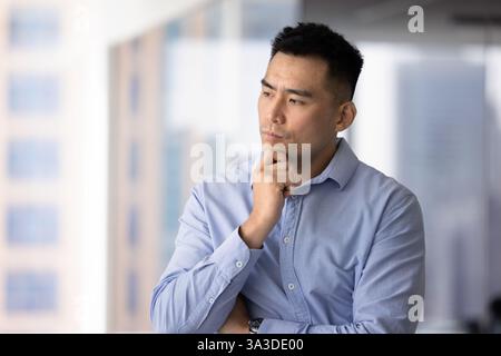 Sérieux et réfléchi bel homme d'affaires asiatique touchant le menton, regardant loin Banque D'Images