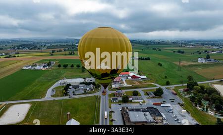 Une montgolfière dynamique flotte gracieusement au-dessus d'un paysage rural pittoresque rempli de champs verts et de bâtiments dispersés. Le ciel nuageux ajoute une atmosphère sereine à la scène. Banque D'Images