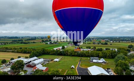 Une montgolfière vibrante survole de vastes champs verdoyants et de structures agricoles sous un ciel nuageux. Le cadre rural offre une toile de fond sereine pour la vue aérienne. Banque D'Images