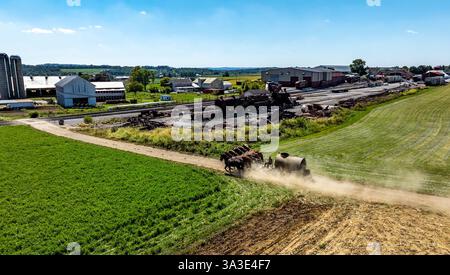 Vue large des terres agricoles avec des chevaux tirant l'équipement de fumier le long d'une route de terre dans le cadre rural, sur une belle journée ensoleillée. Banque D'Images