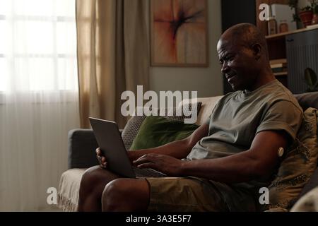 Homme afro-américain assis sur le canapé travaillant sur ordinateur portable. Salon confortable au décor moderne et à la lumière naturelle qui passe par la fenêtre pour améliorer l'ambiance détendue Banque D'Images