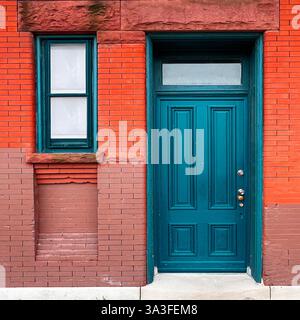 Porte en bois sarcelle avec fenêtre à tableau arrière dans une façade historique en brique rouge et brun, avec textures contrastées et détails architecturaux. Banque D'Images