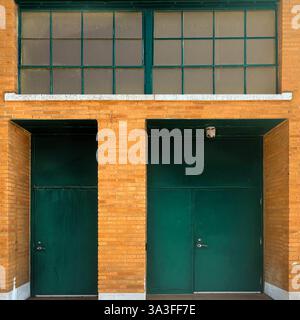 Entrée de bâtiment industriel avec deux portes en métal vert foncé dans une façade en briques jaunes, fenêtres à grille encadrée et un plafonnier de sécurité. Banque D'Images