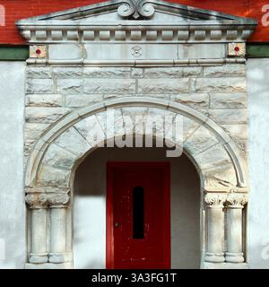 Porte rouge encadrée par une arche en pierre romane ornée avec des détails de colonne complexes, serrée contre une façade historique en briques et en stuc. Banque D'Images