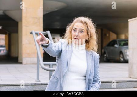 Femme d'affaires mature expressive avec des cheveux blonds bouclés et des lunettes, portant un blazer gris clair et col roulé blanc, pointant vers quelque chose avec un sur Banque D'Images