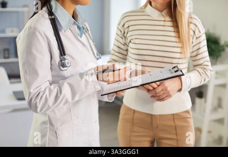 Plan recadré d'une femme médecin en manteau blanc avec presse-papiers discutant des résultats médicaux avec le patient. Banque D'Images