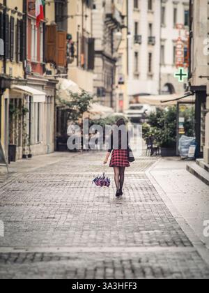 Une jeune femme élégante dans une jupe à carreaux rouge descend une rue pavée italienne tenant un parapluie. L'architecture historique et l'éclairage doux c Banque D'Images