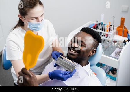 African est à la recherche de facettes ou d'implants de dents de couleur assortie d'échantillons dans les mains du médecin. Odontologie. Dentiste montrant les dents guide de nuances de couleur pour la dent Banque D'Images